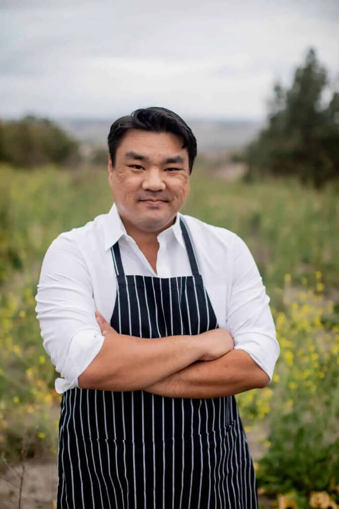 Nathaniel Lee in a chef's apron standing in a field of yellow flowers
